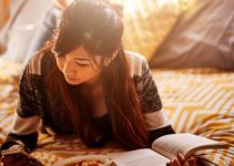 Asian teen on bed checking smartphone while reading book and eating soup;