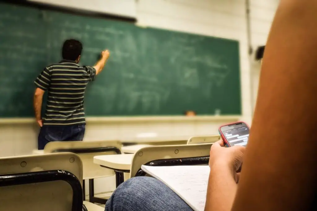 Technology in classroom: Man Writing on Chalkboard While Student Texts on Mobile Phone