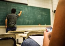 Man Writing on Chalkboard While Student Texts on Mobile Phone