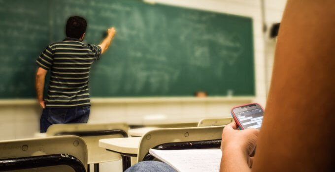 Man Writing on Chalkboard While Student Texts on Mobile Phone